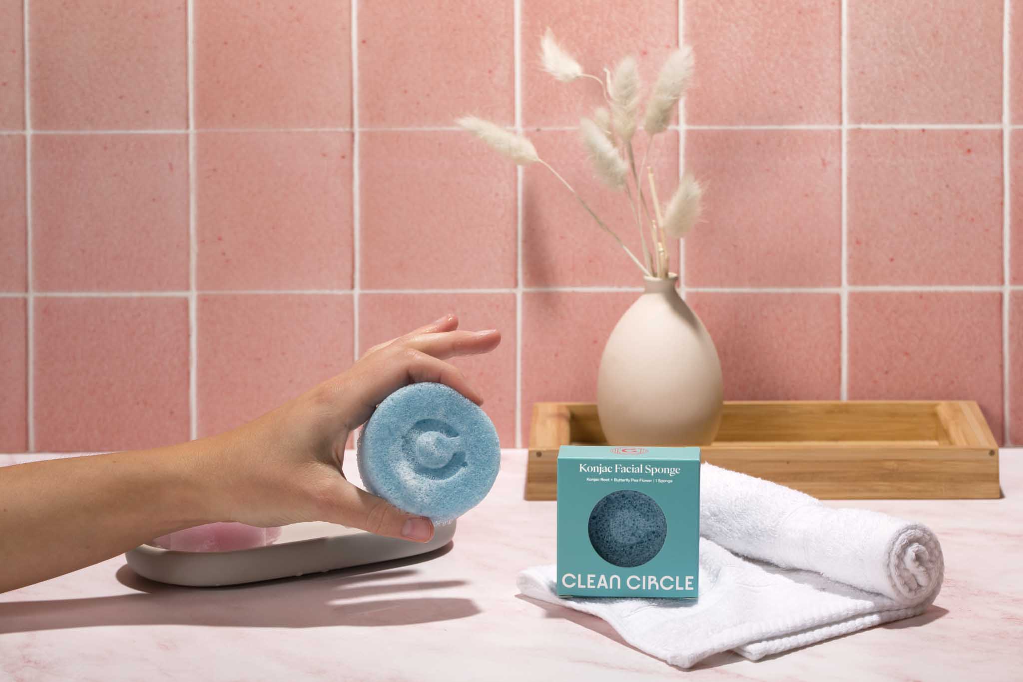 cleancircle- hand holding bluefly peaflower konjac facial sponge in bathroom setting with pink tiles in background and towel on counter